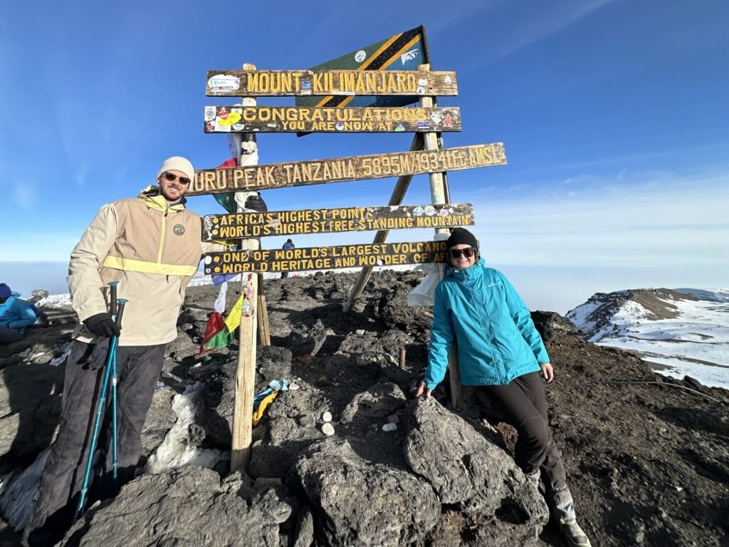 Harry and Renee standing at the summit of Mount Kilimanjaro with stunning clouds and African landscape views